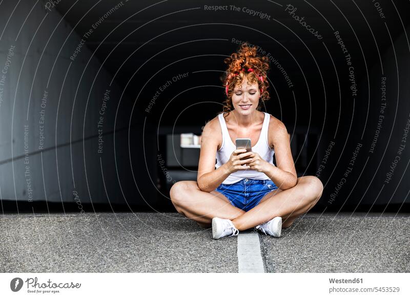 Redheaded woman sitting on parking level holding cell phone females women mobile phone mobiles mobile phones Cellphone cell phones smiling smile Seated Adults