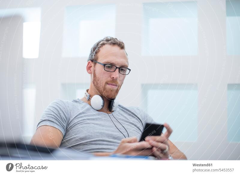 Young man with headphones around his neck reading text messages on his phone cool attitude composed coolness laid-back young man young men Office Offices