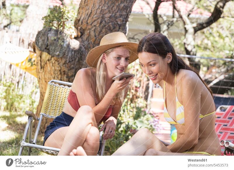 Two young women in garden sharing cell phone female friends mobile phone mobiles mobile phones Cellphone cell phones relaxed relaxation mate friendship