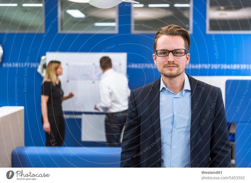 Businessman standing in conference room, while colleagues working with flipchart in background presentation presentations teamwork teamworking Business man