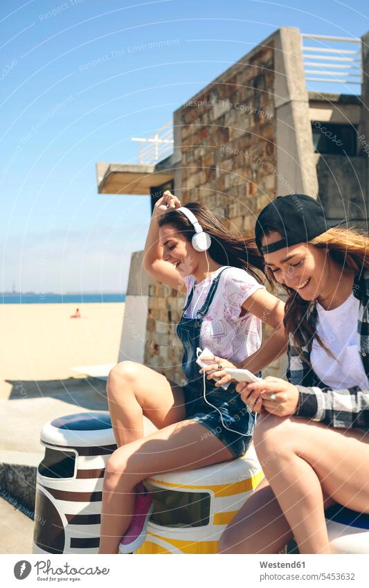 Two happy young women listening music and having fun next to the beach headphones headset sitting Seated hearing Smartphone iPhone Smartphones Listening Music