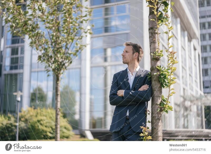 Businessman leaning against a tree in front of office building Business man Businessmen Business men Tree Trees standing office buildings business people