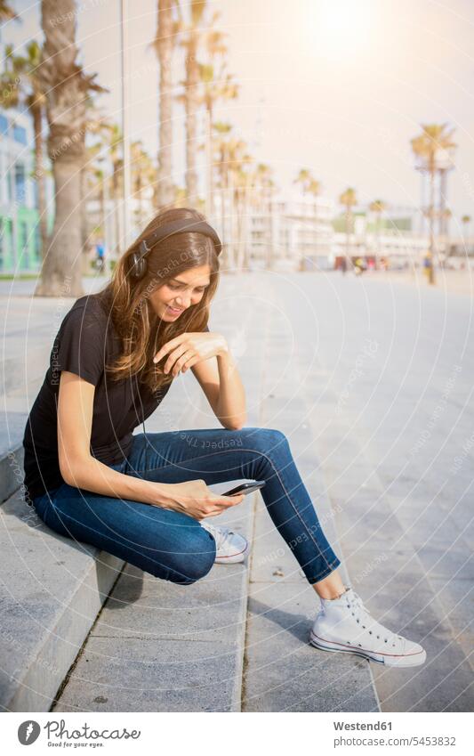 Smiling young woman sitting on steps listening to music smiling smile hearing headphones headset females women Adults grown-ups grownups adult people persons