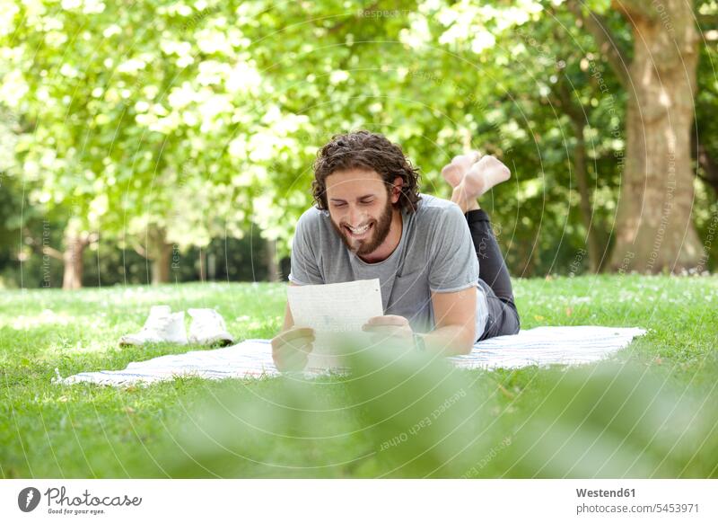 Laughing man lying on blanket in a park reading a letter men males letter document letters Adults grown-ups grownups adult people persons human being humans