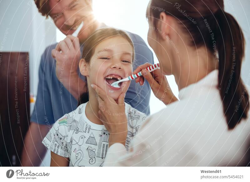 Mother brushing daughter’s teeth in bathroom brushing teeth toothbrush tooth-brushes toothbrushes family families people persons human being humans human beings