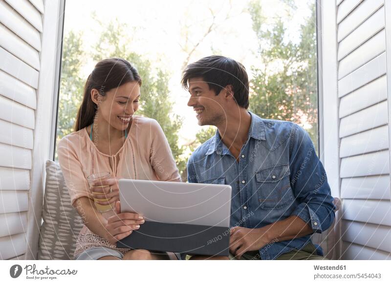 Smiling young couple sitting on windowsill sharing tablet twosomes partnership couples relaxed relaxation digitizer Tablet Computer Tablet PC Tablet Computers