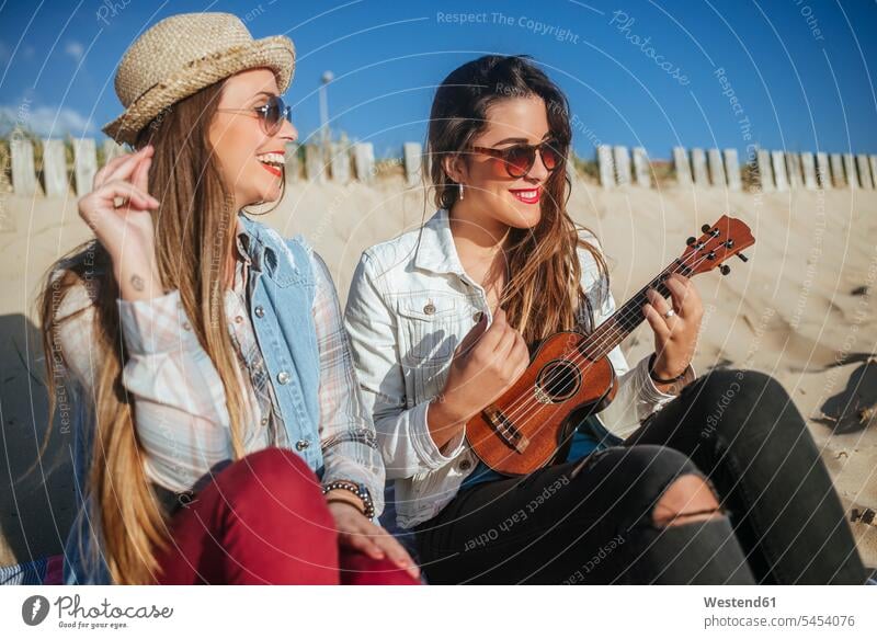 Two young women with ukulele sitting on the beach beaches female friends mate friendship woman females Seated laughing Laughter ukelele sunglasses sun glasses