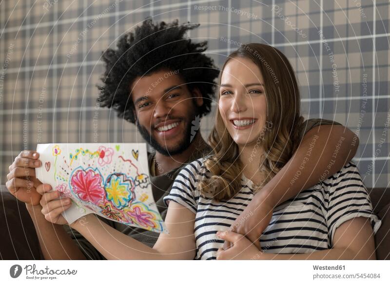 Smiling couple sitting on couch showing children's drawing child's drawing Child's Drawings twosomes partnership couples smiling smile drawings image images