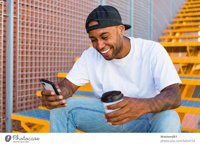 Laughing young man with coffee to go sitting on stairs looking at smartphone laughing Laughter Smartphone iPhone Smartphones men males positive Emotion Feeling
