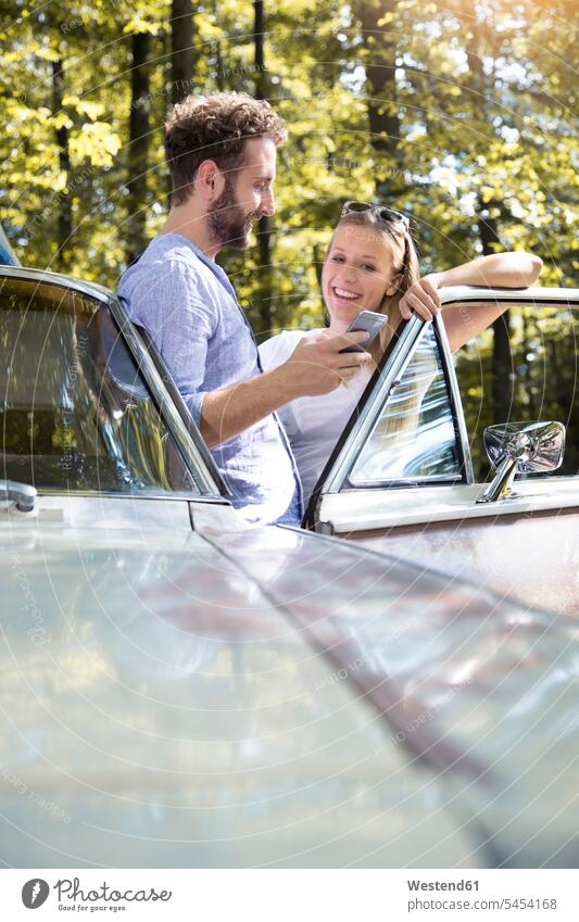 Smiling young couple with cell phone at car in forest smiling smile woods forests mobile phone mobiles mobile phones Cellphone cell phones twosomes partnership
