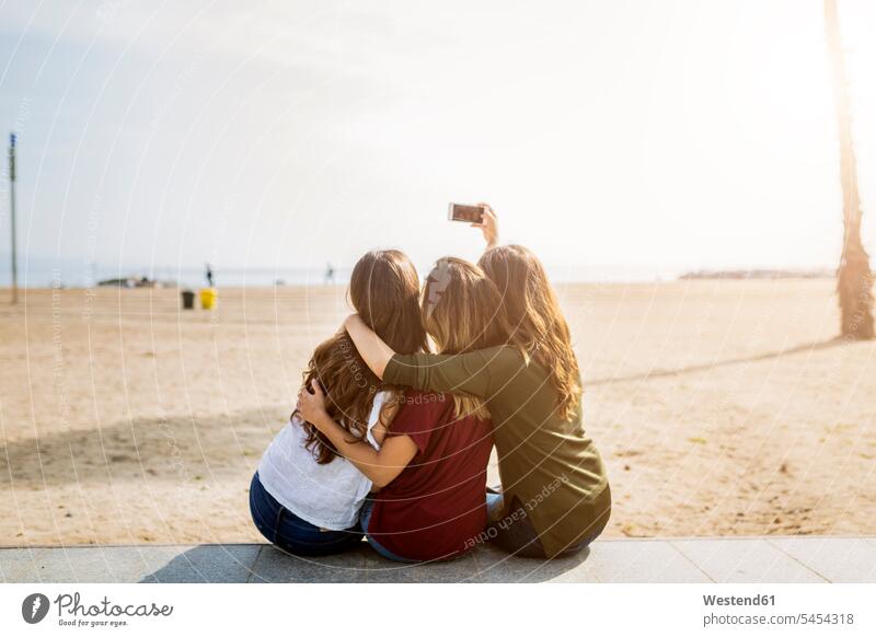 Rear view of three female friends sitting at the beach taking a selfie mobile phone mobiles mobile phones Cellphone cell phone cell phones Selfie Selfies