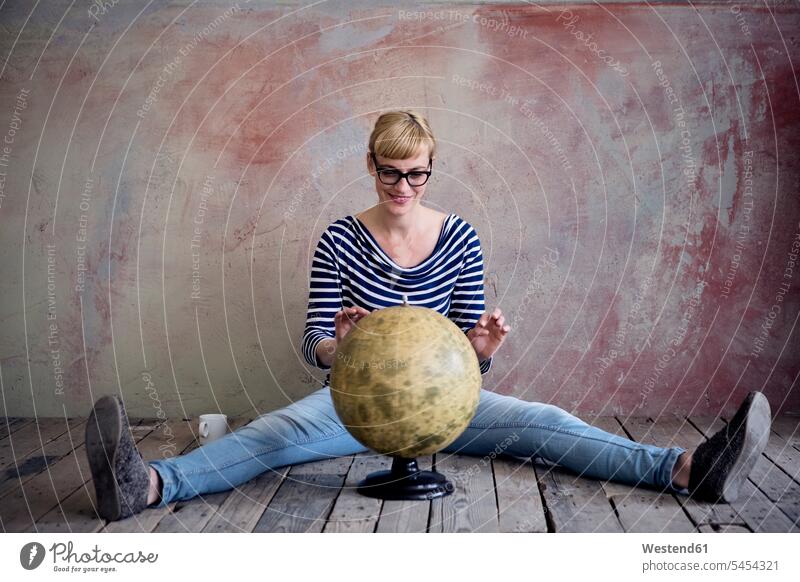 Smiling woman sitting on wooden floor in an unrenovated room looking at globe globes females women Adults grown-ups grownups adult people persons human being