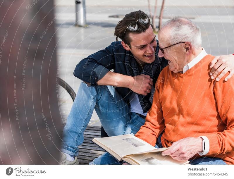 Happy senior man and adult grandson with photo album outdoors grandsons looking eyeing smiling smile bench benches happiness happy grandfather grandpas