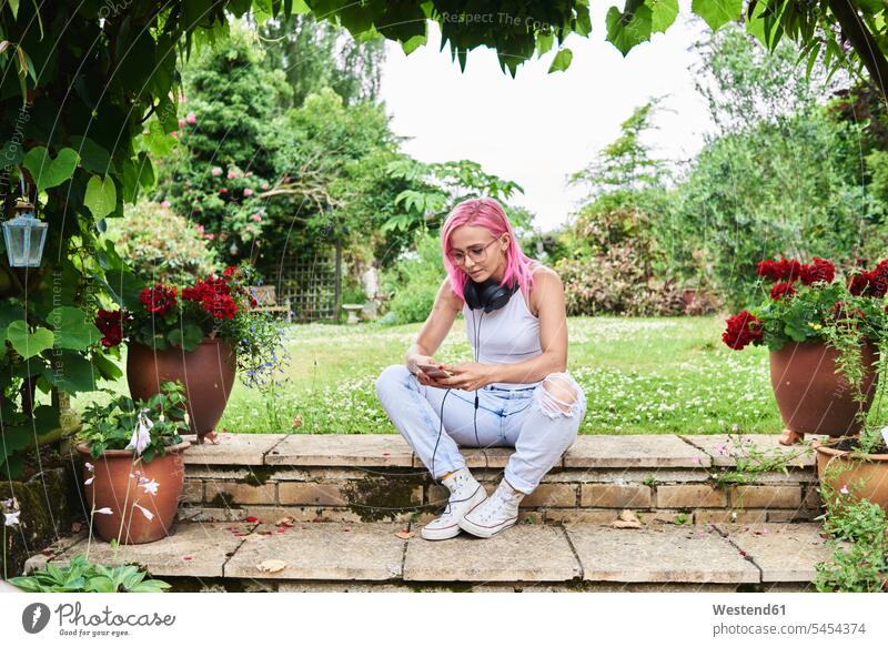 Young woman with pink hair wearing headphones and using cell phone in garden headset sitting Seated females women mobile phone mobiles mobile phones Cellphone