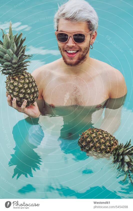 Smiling young man holding pineapples in swiming pool Pineapple Ananas comosus Ananas sativus Pineapples swimming pool pools swimming pools men males Fruit