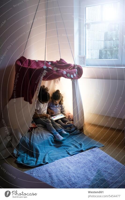 Two sisters sitting in dark children's room, looking at digital tablet playing Kids Room nursery child's room reading Seated rooms domestic room domestic rooms