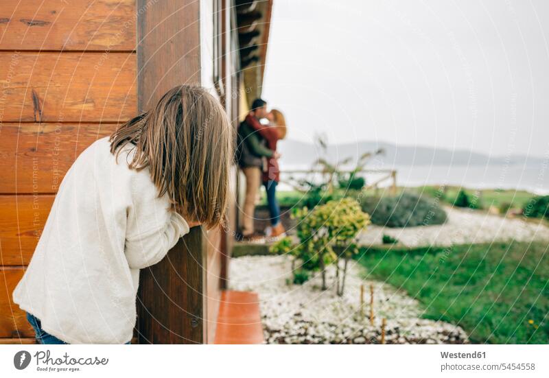 Back view of little girl spying couple in love kissing next to wooden house parents watching observing observe family families people persons human being humans