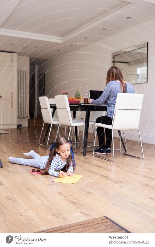 Little girl lying on floor drawing a picture while her mother working on laptop in the background females girls child children kid kids people persons
