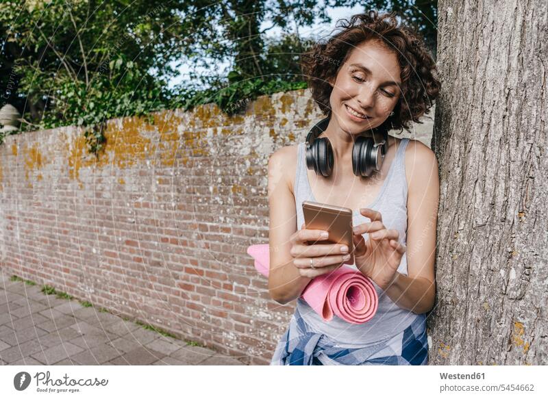 Smiling woman with mat, headphones and cell phone on pavement mobile phone mobiles mobile phones Cellphone cell phones females women telephones communication
