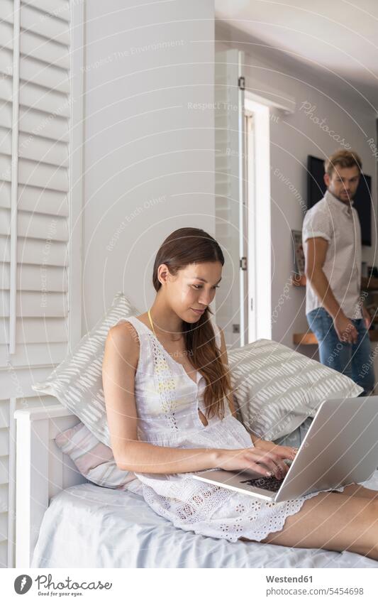 Young woman using laptop in bedroom with man in background Laptop Computers laptops notebook couple twosomes partnership couples smiling smile computer
