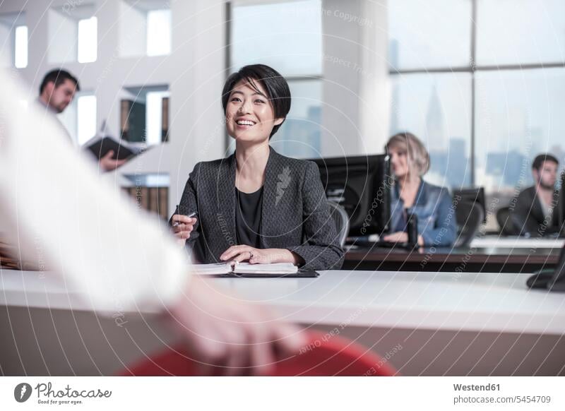 Smiling woman sitting at desk in city office smiling smile talking speaking offices office room office rooms notebook notebooks colleagues workplace work place