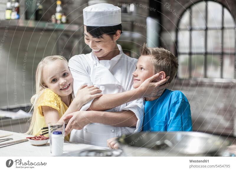 Female chef with happy boy and girl in cooking class kitchen female cook cooks Chefs instruction tuition looking at camera looking to camera