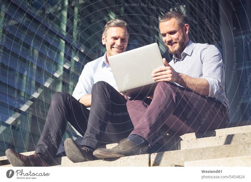 Two smiling businessmen sitting on stairs using laptop stairway Laptop Computers laptops notebook Seated colleagues Businessman Business man Businessmen