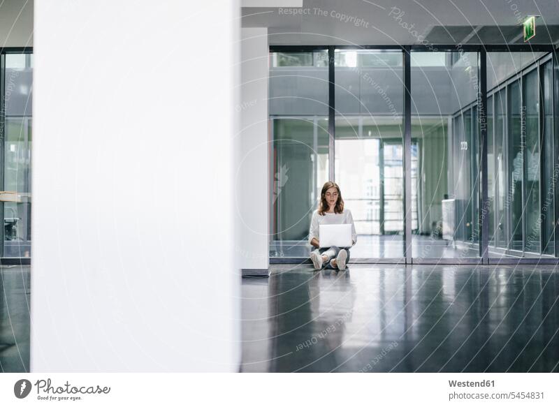 Businesswoman sitting on ground in empty office, using laptop offices office room office rooms Sitting On The Floor Sitting On Floor Seated working At Work