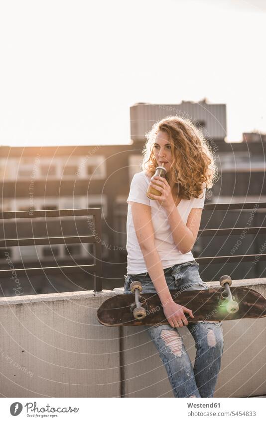 Young woman with skateboard drinking beverage on roof terrace at sunset sunsets sundown Skate Board skateboards females women Drink beverages Drinks Beverage