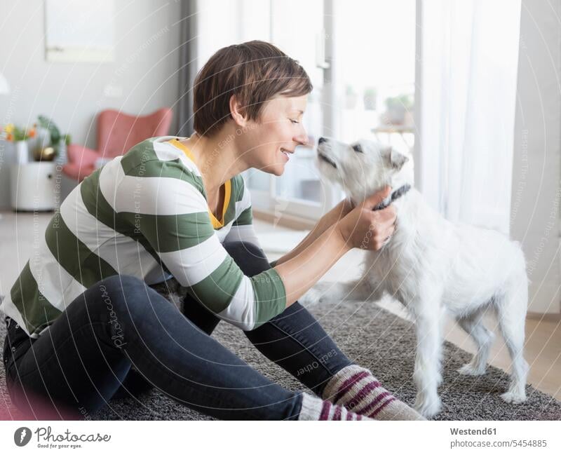 Smiling woman sitting on the floor in the living room cuddling with her dog dogs Canine females women pets animal creatures animals Adults grown-ups grownups