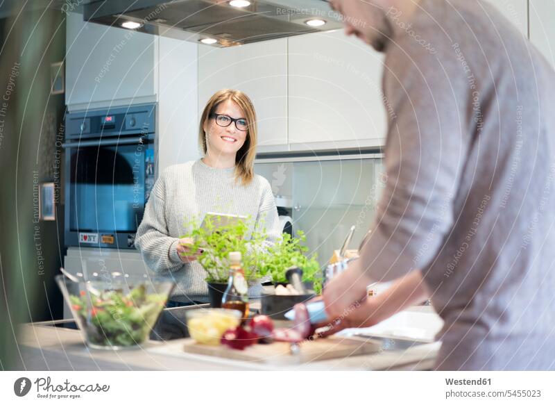 Smiling woman with tablet looking at boyfriend preparing salad in kitchen couple twosomes partnership couples smiling smile digitizer Tablet Computer Tablet PC