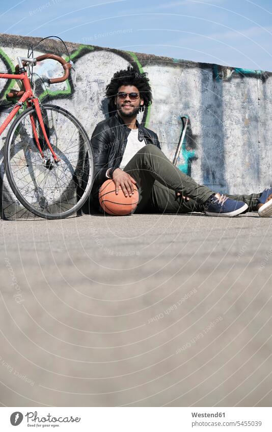 Smiling man sitting in front of grafitti wall next to his bicycle holding basketball men males smiling smile bikes bicycles Seated Adults grown-ups grownups