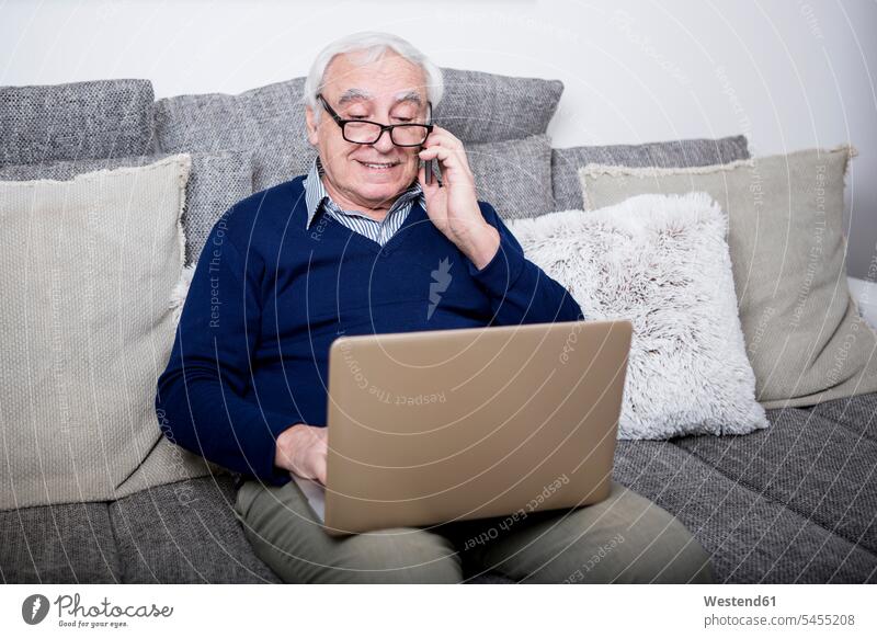 Senior man sitting on couch, using laptop and smartphone settee sofa sofas couches settees senior men senior man elder man elder men senior citizen on the phone