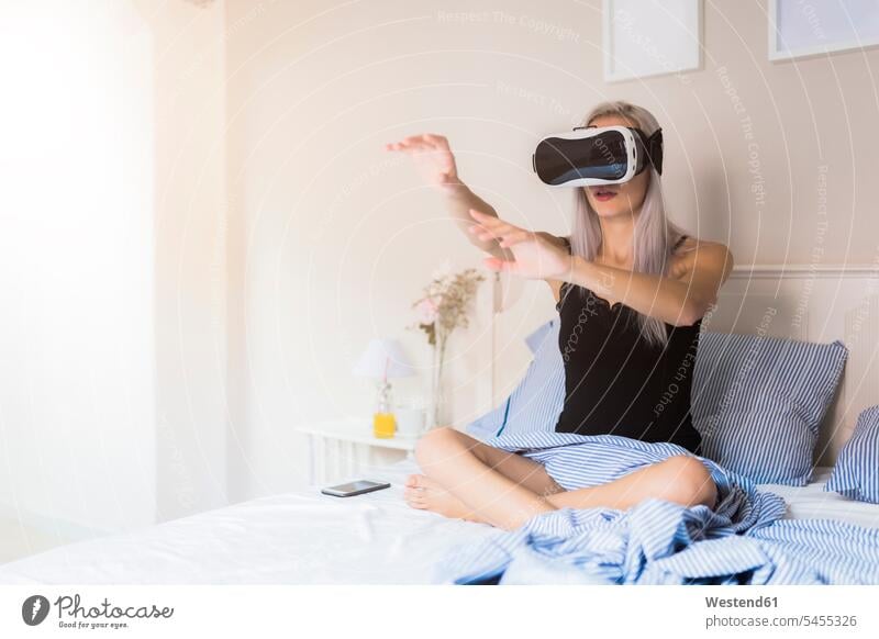 Young woman sitting in bed wearing VR glasses virtual reality specs Eye Glasses spectacles Eyeglasses beds females women Seated Adults grown-ups grownups adult