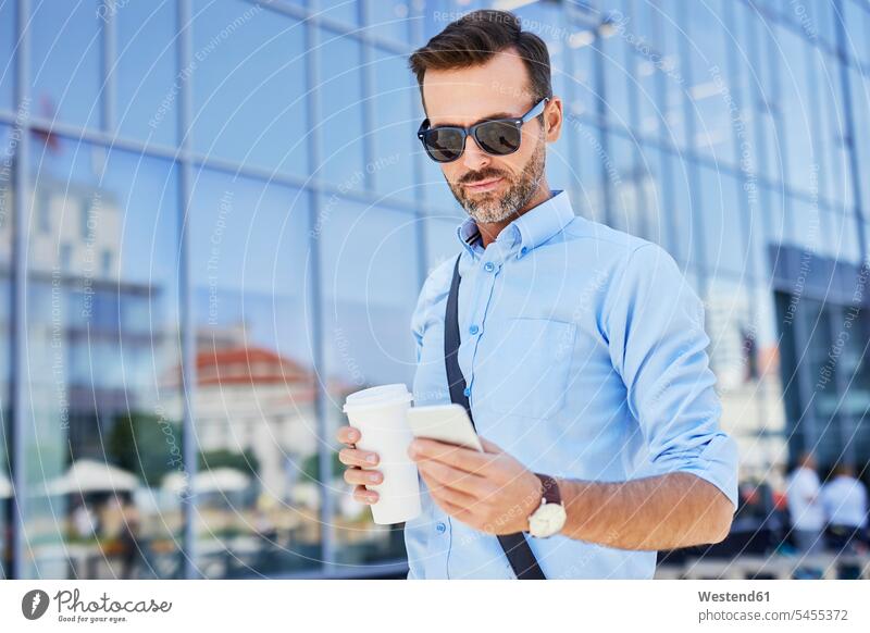 Businessman using phone and holding coffee outdoors Coffee standing mobile phone mobiles mobile phones Cellphone cell phone cell phones Business man Businessmen