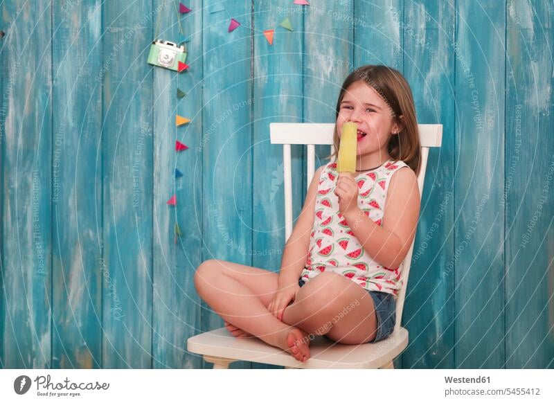 Happy little girl sitting on chair eating lemon ice lolly Seated chairs females girls Sweet Food sweet foods food and drink Nutrition Alimentation