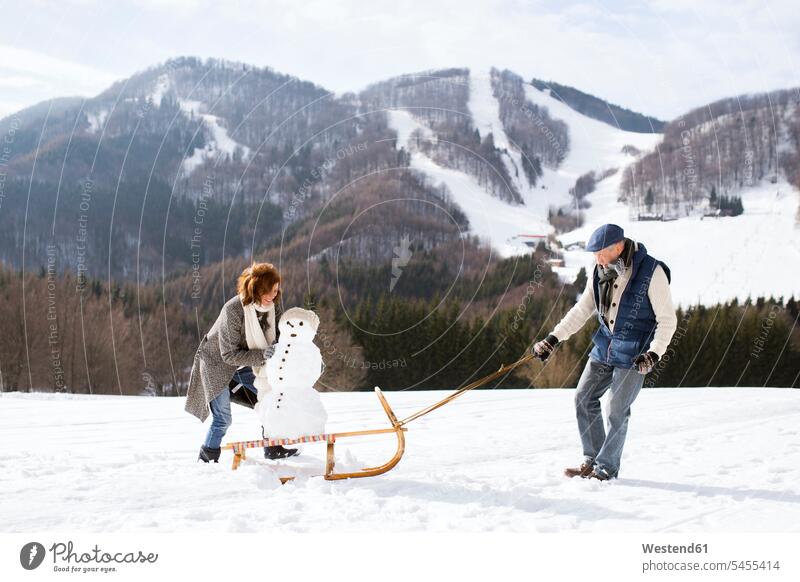 Senior couple with snowman on sledge in winter landscape snowmen senior couple elder couples senior couples adult couple adult couples twosomes partnership
