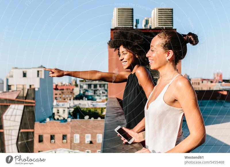 Female friends standing on rooftop, pointing at distance smiling smile Joy enjoyment pleasure Pleasant delight roof terrace deck female friends Smartphone