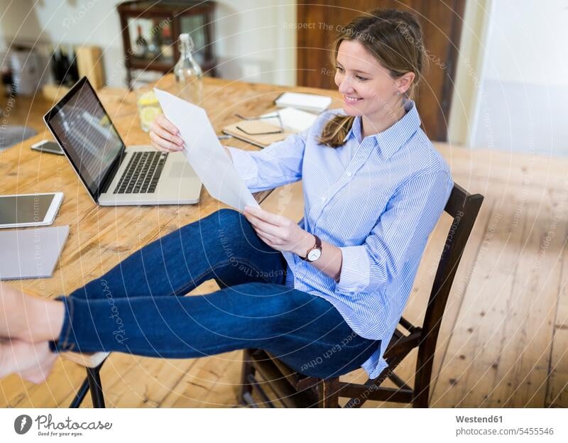 Smiling woman sitting at desk at home with feet up reading document Seated females women documents desks foot human foot human feet Adults grown-ups grownups
