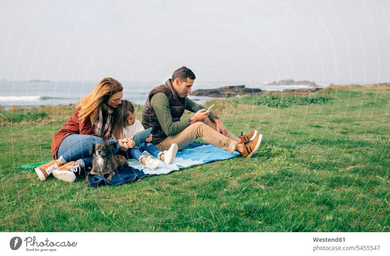 Family with dog sitting on blanket at the coast using wireless devices dogs Canine coastline shoreline Blanket Blankets family families pets animal creatures