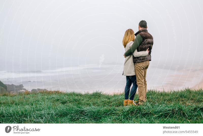Couple hugging at the coast on a foggy winter day looking at view embracing embrace Embracement couple twosomes partnership couples coastline shoreline people