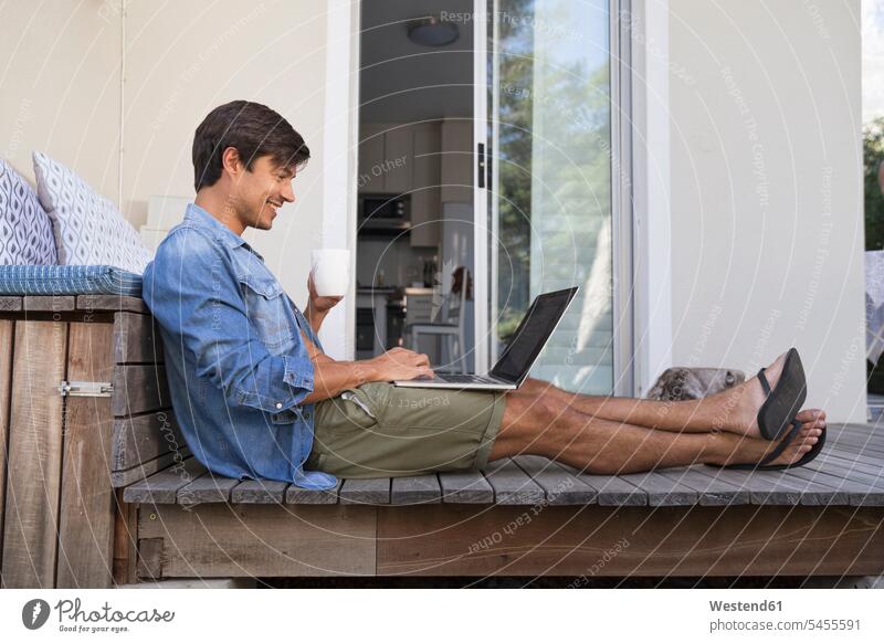 Man sitting on terrace using laptop Laptop Computers laptops notebook smiling smile terraces man men males Seated computer computers Adults grown-ups grownups