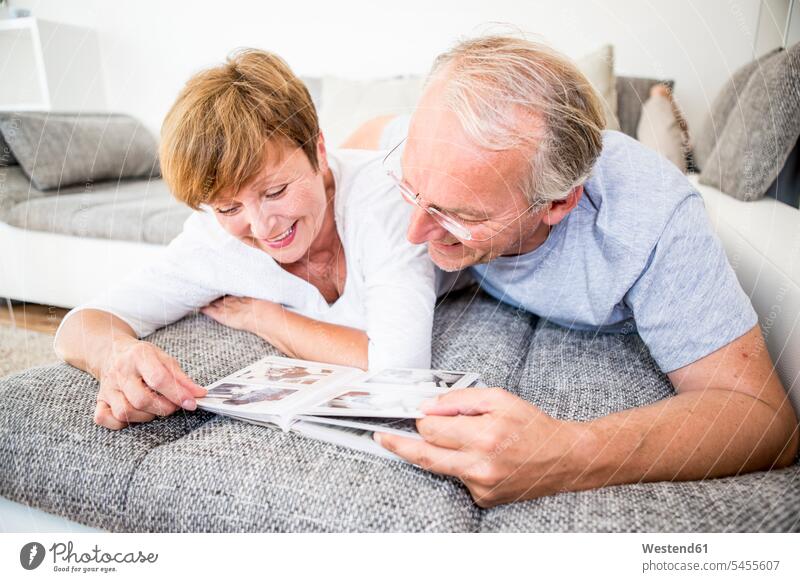 Senior couple at home lying on couch looking at photo album photograph album photo albums settee sofa sofas couches settees smiling smile relaxed relaxation
