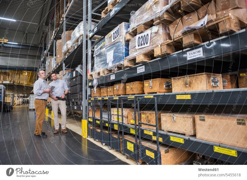 Two men standing in factory warehouse talking speaking colleagues factories man males Adults grown-ups grownups adult people persons human being humans