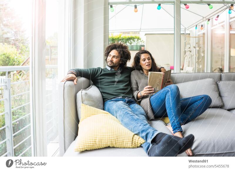 Couple sitting on couch reading book Seated settee sofa sofas couches settees couple twosomes partnership couples people persons human being humans human beings
