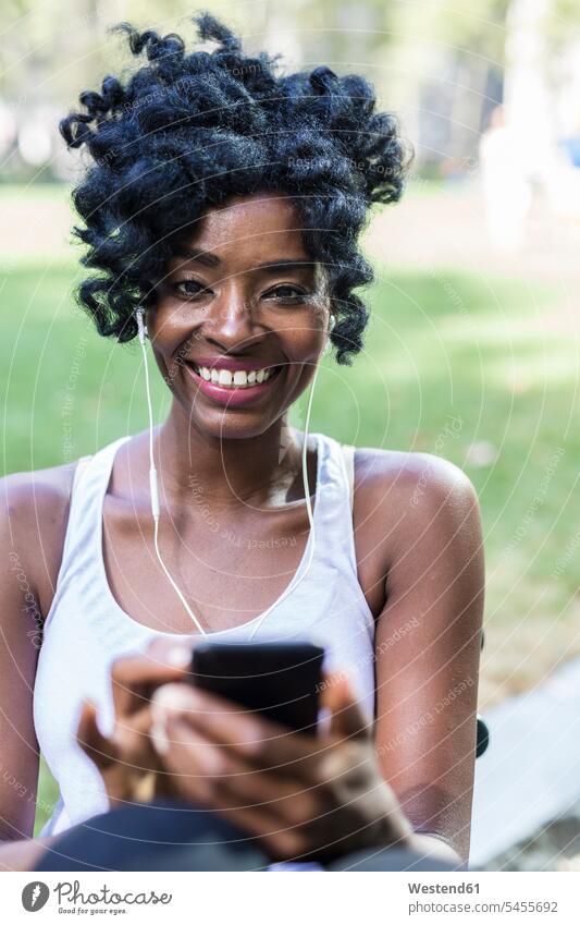 Portrait od smiling woman using cell phone and earphones in a park females women Smartphone iPhone Smartphones portrait portraits Adults grown-ups grownups