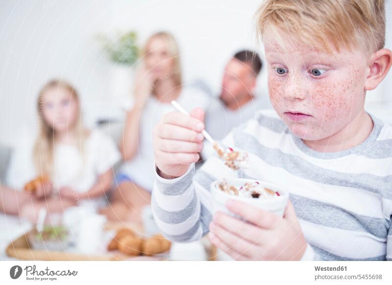 Boy staring at cereal bowl with family in background eating boy boys males child children kid kids people persons human being humans human beings families home