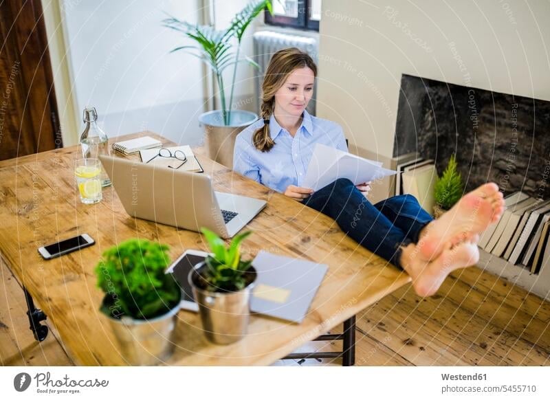 Woman sitting at desk at home with feet up reading document woman females women foot human foot human feet Seated documents desks Adults grown-ups grownups