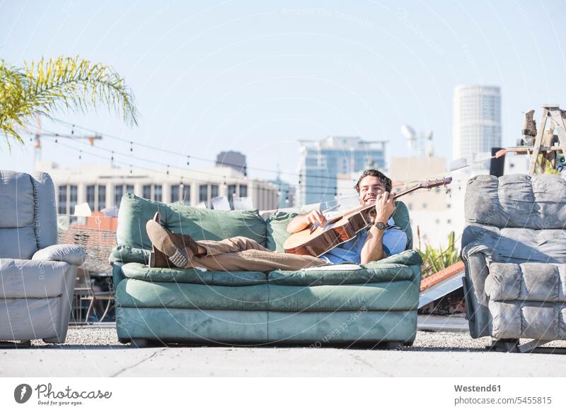 Young man on rooftop lying on sofa and playing guitar musician musicians couch settee sofas couches settees guitars roof terrace deck cheerful gaiety Joyous