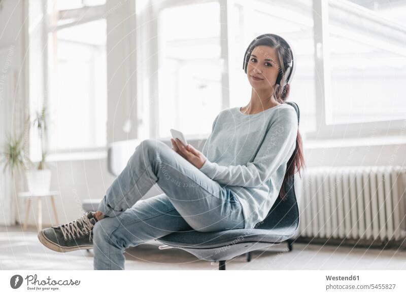 Young woman making a video call from her mobile phone, wearing headphones females women headset chair chairs mobiles mobile phones Cellphone cell phone